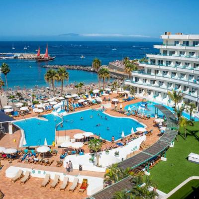 Aerial view of a seaside resort with a large swimming pool, sun loungers and a white multi-storey hotel beside a beach and marina in Tenerife