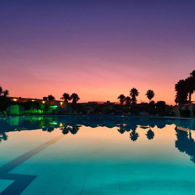 Sunset over a resort swimming pool with palm trees silhouetted against a purple and orange sky, reflected in the water