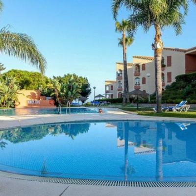 Outdoor swimming pool with palm trees, sun loungers and a Spanish-style resort apartment building under a clear blue sky
