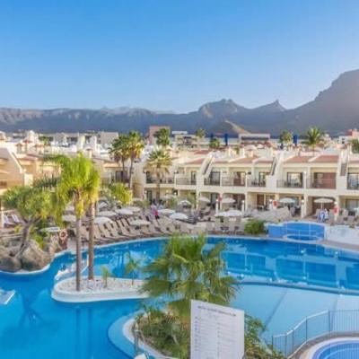 Sunlit resort complex with a large swimming pool, sun loungers, palm trees and white apartment buildings, with mountains in the background