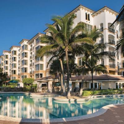 Outdoor swimming pool with sun loungers and palm trees in front of a multi-storey resort building under a clear blue sky
