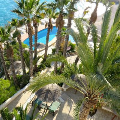 Overhead view of a seaside resort pool area with palm trees, sun loungers and a thatched parasol beside turquoise water