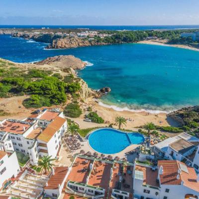 Aerial view of a Spanish coastal resort with terracotta-roof buildings, a large swimming pool and a sandy bay with turquoise sea and rocky headland