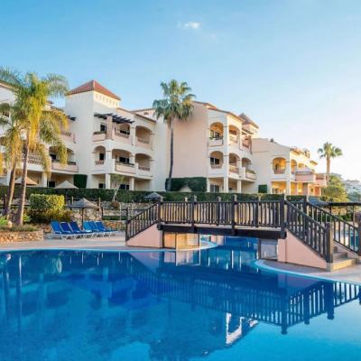 Outdoor swimming pool with a small wooden bridge, sun loungers and palm trees in front of a white resort building under a clear blue sky
