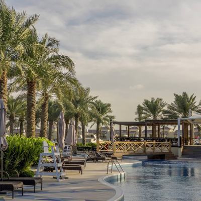Palm-lined resort pool area with sun loungers, closed parasols and a wooden terrace under a cloudy sky