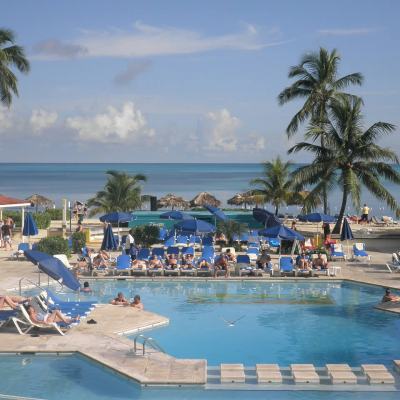 Busy beachfront resort swimming pool with sun loungers, blue parasols and palm trees overlooking the sea