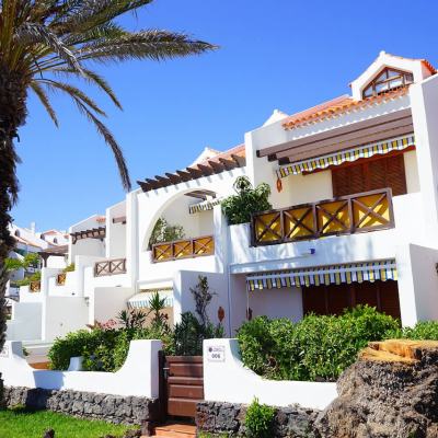 White Mediterranean-style holiday apartments with striped awnings and balconies, framed by a large palm tree under a clear blue sky