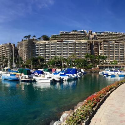 Marina with moored yachts beside a waterfront promenade, overlooking a large terraced resort complex on a hillside under a clear blue sky