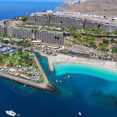 Aerial view of a large seaside resort complex with hotel buildings, marina, palm-lined pools and a sandy beach beside turquoise water and rocky coastline