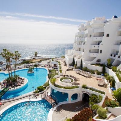 Seafront resort with white terraced apartments overlooking a turquoise swimming pool, sun loungers and palm trees by the ocean