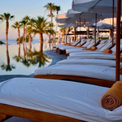 Sun loungers with white cushions and rolled towels beside a resort swimming pool lined with palm trees at sunset