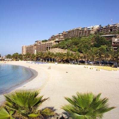Wide view of a sandy beach with calm sea, palm trees and terraced resort buildings on a hillside under a clear blue sky