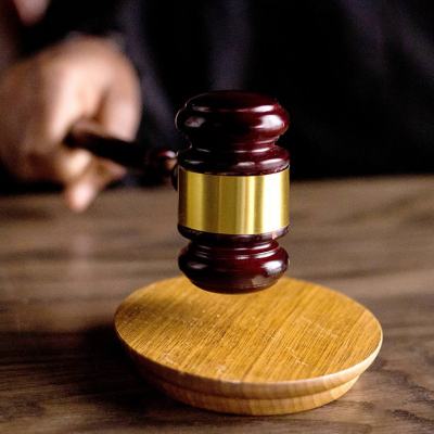 Close-up of a judge’s gavel above a wooden sound block on a desk, with a blurred hand in the background