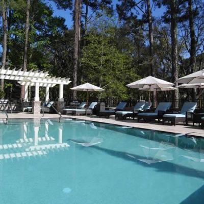 Outdoor swimming pool with sun loungers and parasols beside a white pergola, surrounded by tall pine trees