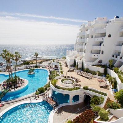 White seaside resort with tiered balconies overlooking sunlit outdoor swimming pools by the ocean, with palm trees and sun loungers on the poolside terrace