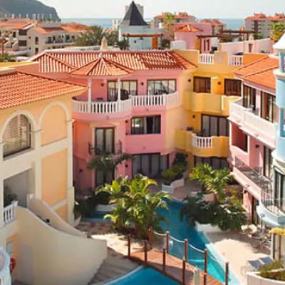 Colourful Mediterranean-style resort buildings with balconies and a turquoise pool, with the sea in the distance