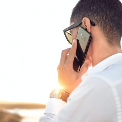 Man in a white shirt holding a smartphone to his ear outdoors, back view with bright sunlight in the background