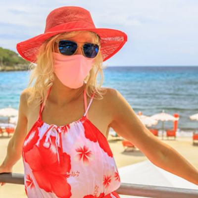 Woman in red sunhat and face mask standing by a railing with a beach and parasols in the background