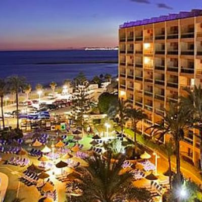 Seafront resort hotel at dusk with illuminated balconies, swimming pool, palm trees and sun loungers beside the shoreline and sea