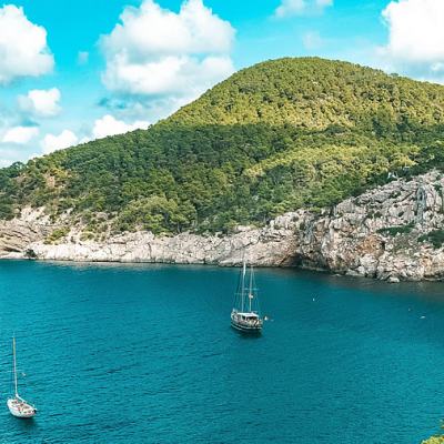 Sailboats anchored in a turquoise bay below rocky cliffs and green hills under a blue sky with scattered clouds