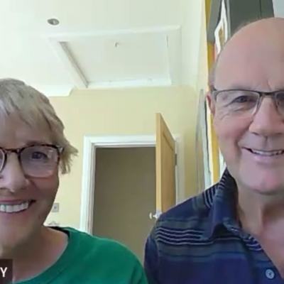 Douglas and Linda Mackay smiling at the camera during a video call, seated indoors at home with an open door behind them