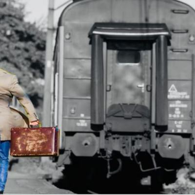 Man with suitcase running alongside a departing train on a railway platform, waving as it pulls away
