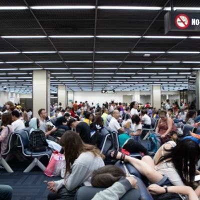Crowded airport departure lounge with passengers sitting on the floor and seats beneath a “No-smoking area” sign during flight cancellations