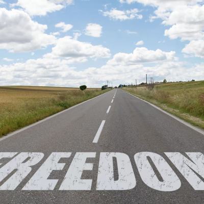 Long straight country road under a cloudy blue sky with the word “FREEDOM” painted across the tarmac in large white letters