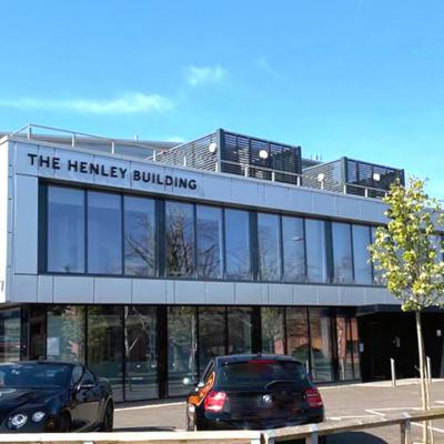 Exterior of The Henley Building office block with large glass windows, parked cars and young trees in front under a blue sky