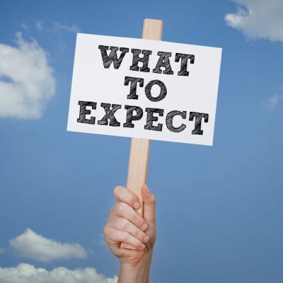 Hand holding a wooden sign reading “What to expect” against a blue sky with clouds