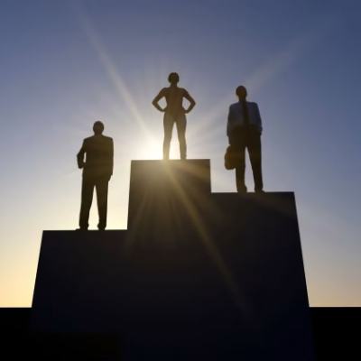Silhouettes of three businesspeople standing on winners podium steps against a sunburst sky at sunset