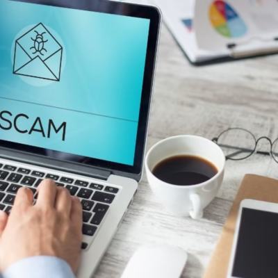 Laptop screen showing an envelope icon and the word SCAM on a desk with hands typing, a coffee cup, glasses, phone and notepad