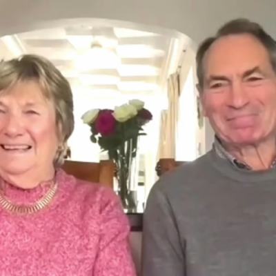 Smiling older couple sitting indoors in a bright room, with a vase of flowers behind them during a video call interview about a timeshare claims win story