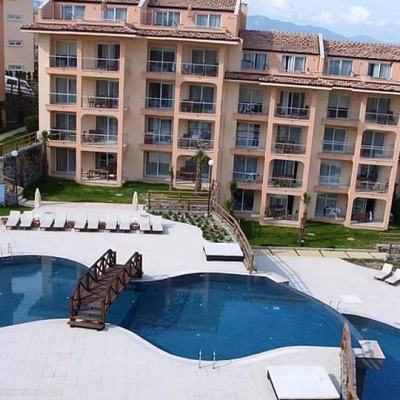 Large apartment-style resort complex with an outdoor swimming pool, sun loungers and parasols on a terrace with hills in the background