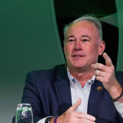 Man in a suit speaking and gesturing at a table with water bottles and a takeaway coffee cup against a green background