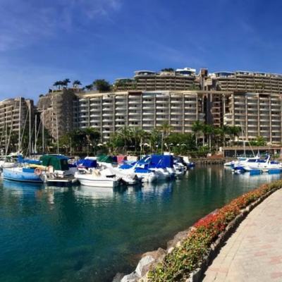 Marina with moored yachts beside a waterfront promenade, with a large terraced resort complex on the hillside in Gran Canaria under a clear blue sky