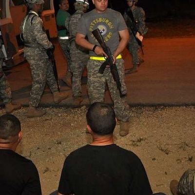 Soldiers with rifles stand guard beside detained men sitting on the ground at night near a vehicle in Mexico