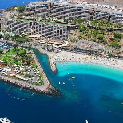 Aerial view of a large coastal resort with multi-storey hotel buildings, marina, palm-lined grounds and a crowded sandy beach beside a turquoise bay
