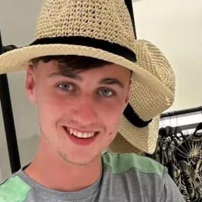 Young man smiling in a straw hat inside a clothing shop, with hanging hats and patterned garments behind him