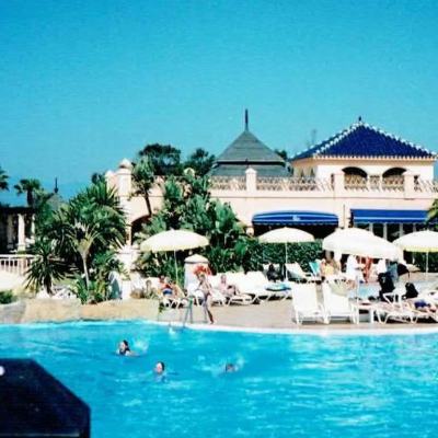 Sunlit resort swimming pool with people relaxing on loungers and parasols beside a Mediterranean-style hotel building and palm trees by the sea