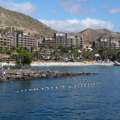 Coastal holiday resort apartments on a hillside behind a sandy beach and calm bay with palm trees and sunloungers