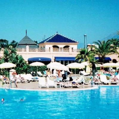 Outdoor hotel pool with sun loungers and parasols in front of a Spanish-style resort building with palm trees and mountains in the background